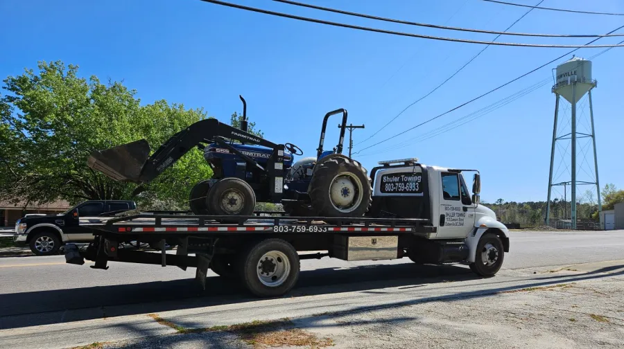 A white Shuler Towing truck with 803-759-6933 on the side carries a blue Farmtrac 555 tractor with a front loader.
